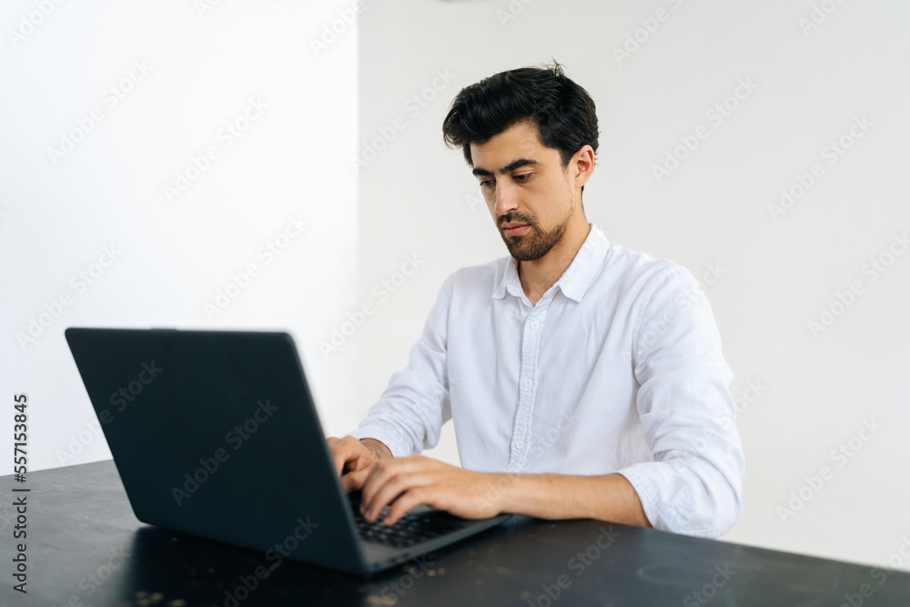 Studio shot of pensive young Caucasian man working online on laptop sitting at table in room with light interior. Thoughtful bearded male looking at computer screen browsing wireless internet. Studio shot of pensive young Caucasian man working online on laptop sitting at table in room with light interior. Thoughtful bearded male looking at computer screen browsing wireless internet.