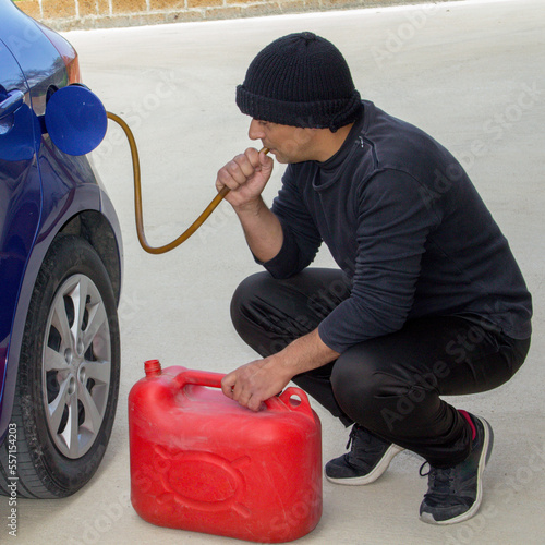 Fotografie Image of a thief dressed in black who sucks petrol from a car tank with a rubber hose