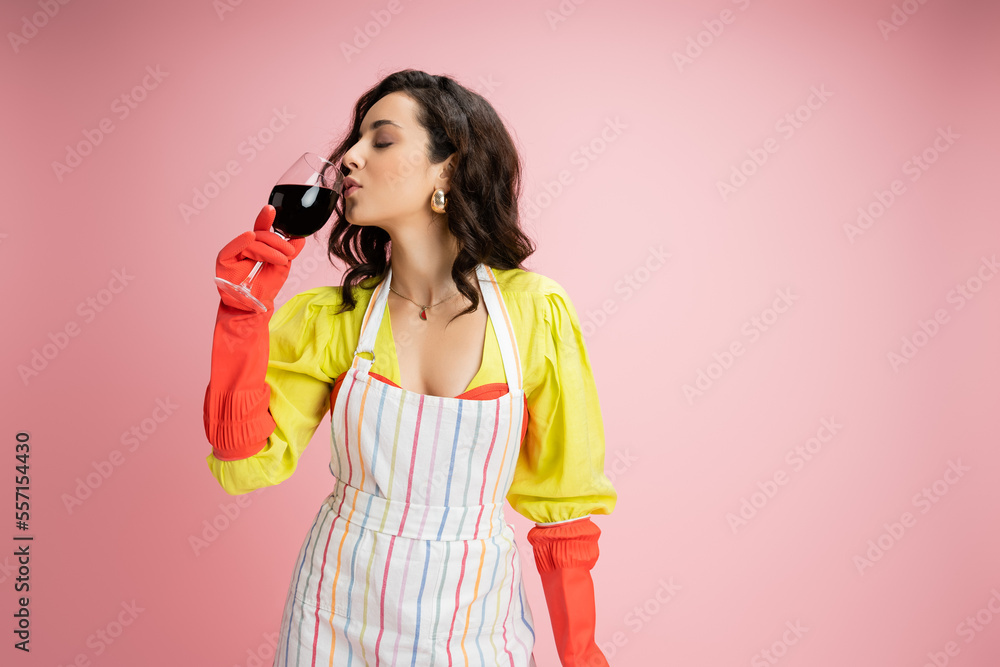 brunette housewife in yellow blouse and striped apron with red rubber gloves drinking wine isolated on pink brunette housewife in yellow blouse and striped apron with red rubber gloves drinking wine isolated on pink