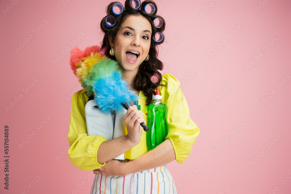 astonished housewife in hair curlers holding detergents and bright feather duster isolated on pink astonished housewife in hair curlers holding detergents and bright feather duster isolated on pink