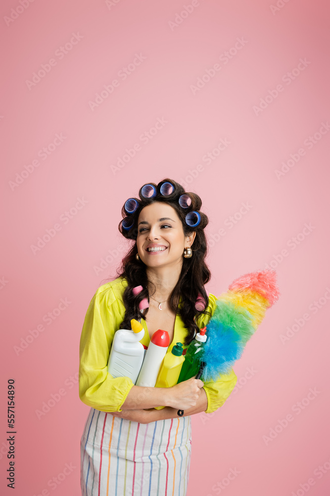 pretty housewife with hair curlers and cleaning supplies smiling and looking away isolated on pink pretty housewife with hair curlers and cleaning supplies smiling and looking away isolated on pink