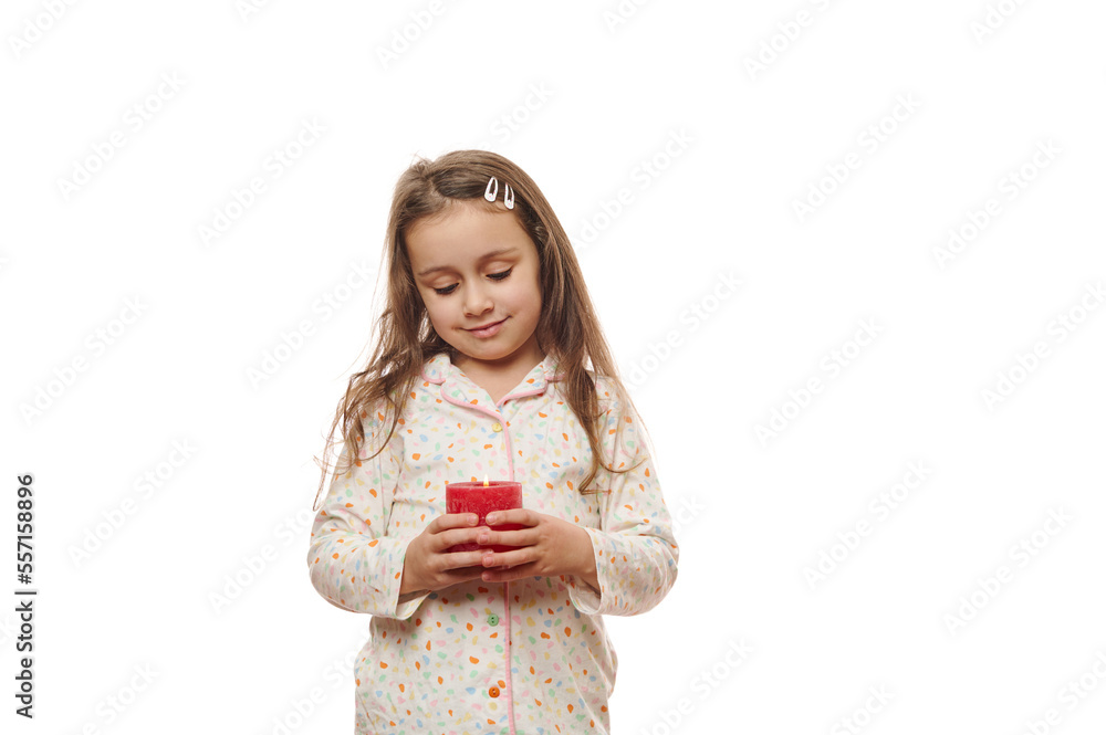 Isolated portrait on white background of a beautiful Caucasian child 5-6 years old, a lovely little girl in comfortable soft pajamas, holding a lit red candle, before going to bed. Copy ad space Isolated portrait on white background of a beautiful Caucasian child 5-6 years old, a lovely little girl in comfortable soft pajamas, holding a lit red candle, before going to bed. Copy ad space