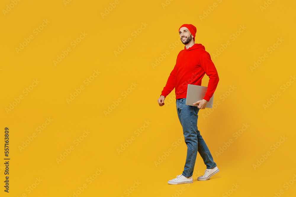 Full body side view smiling fun young caucasian IT man wear red hoody hat look camera hold closed laptop pc computer isolated on plain yellow color background studio portrait People lifestyle concept Full body side view smiling fun young caucasian IT man wear red hoody hat look camera hold closed laptop pc computer isolated on plain yellow color background studio portrait People lifestyle concept