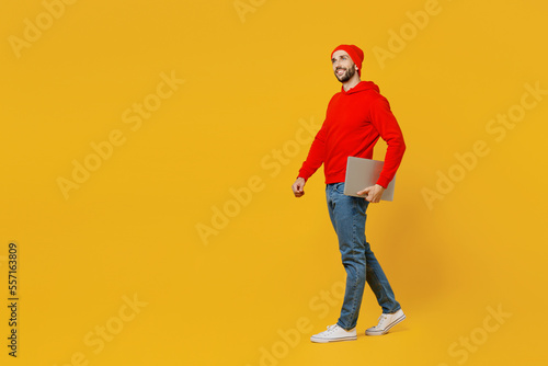 Full body side view smiling fun young caucasian IT man wear red hoody hat look camera hold closed laptop pc computer isolated on plain yellow color background studio portrait People lifestyle concept