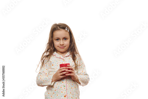 Adorable sleepy baby girl in stylish soft pajamas with colorful dots, cutely smiling at camera, posing with red lit candle in her hands, isolated over white background. Copy advertising space for text