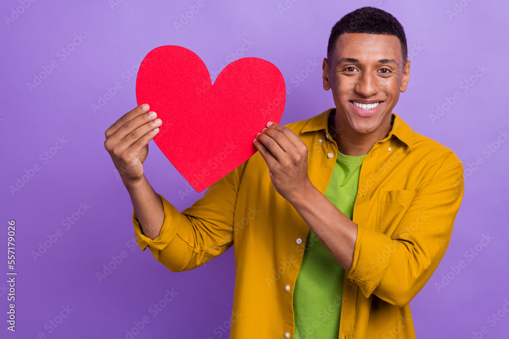 Photo of funny adorable man wear yellow shirt rising large red heart smiling isolated violet color background Photo of funny adorable man wear yellow shirt rising large red heart smiling isolated violet color background