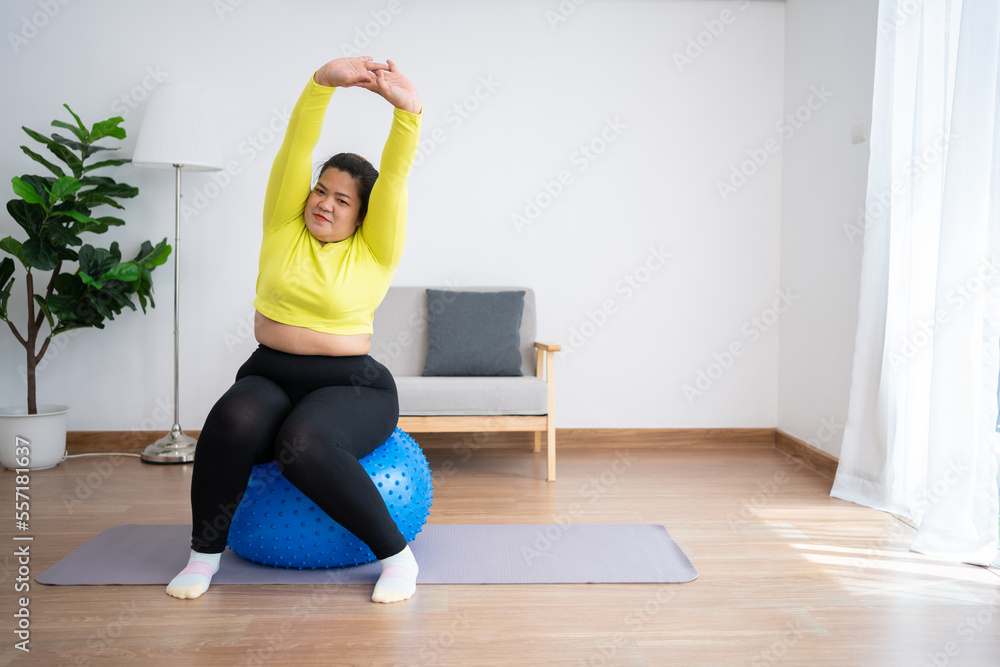 Portrait plus size woman doing exercise with fitness ball in home gym. Overweight woman sitting on a pilates ball and Stretching her muscles before exercising. Health care and weight loss concept.