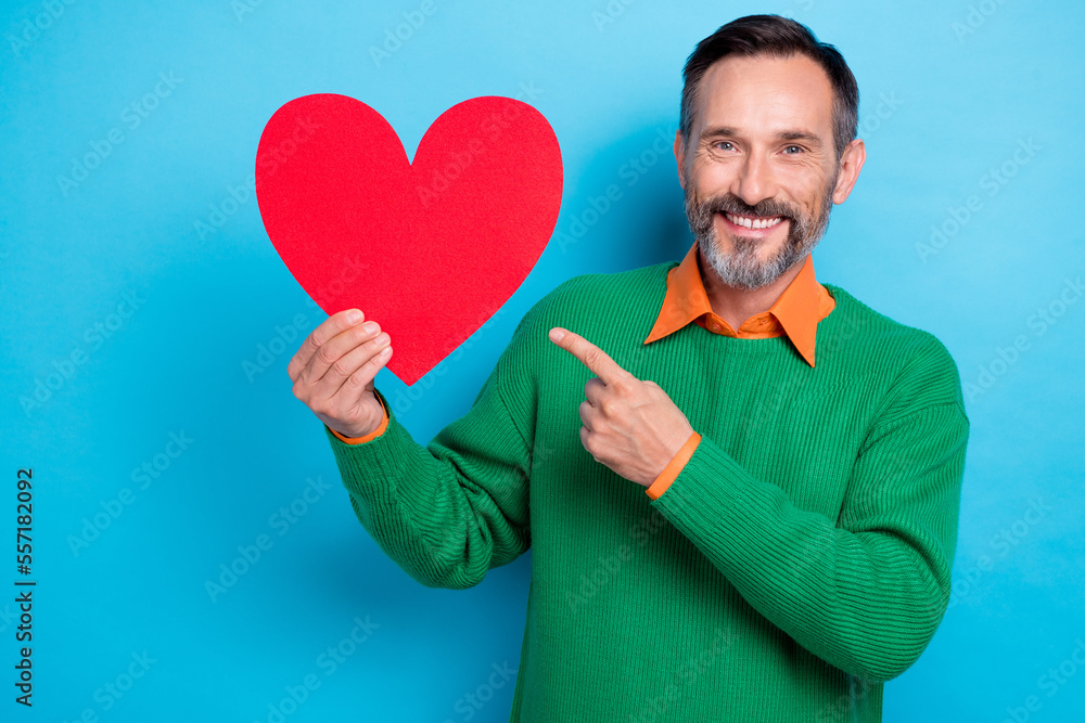 Photo portrait of mature nice mister hold point red heart shape donation promo wear trendy green outfit isolated on blue color background Photo portrait of mature nice mister hold point red heart shape donation promo wear trendy green outfit isolated on blue color background