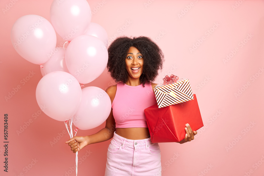 Cheerful positive young curly haired female model looks excited, holds presents and bunch of balloons, celebrated birthday, stands againsta pink background Cheerful positive young curly haired female model looks excited, holds presents and bunch of balloons, celebrated birthday, stands againsta pink background