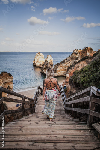 a tattooed woman in a dress walking down wooden stairs leading to a beautiful cliff sand beach strand in Portugal