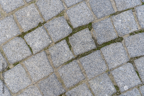 Natural stone background and texture in gray color close up. Pavement paved with square and uneven stones. Paving street with pattern. Between crushed cobblestones there are earth sand and grass.