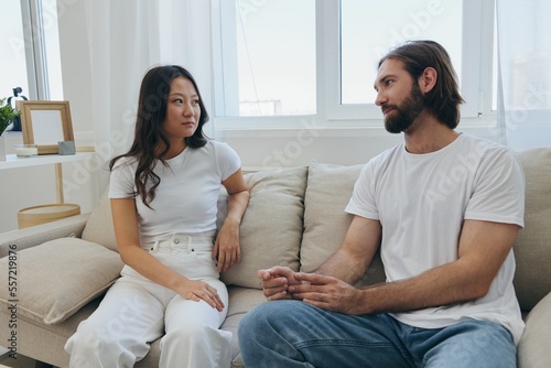 A man and a woman sitting at home on the couch in white stylish t-shirts and chatting merrily smiling and laughing at home. Male and female friendship