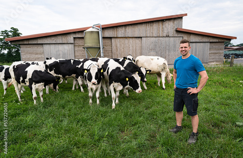 Junger Landwirt steht mit seinen Rindern auf einer Wiese, die Tiere kommen langsam näher, im Hintergrund ein Rinderstall. photo