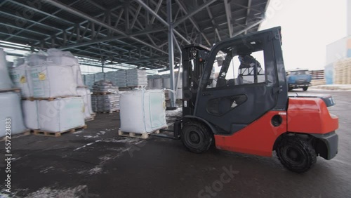 Worker lifts and moves packages of building materials by electric forklift on winter day. Warehouse with modern polycarbonate panels and coils of film