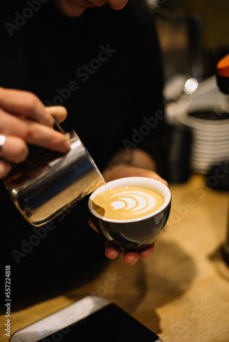 Young male barista pouring frothed milk into the cup with freshly brewed double espresso coffee, making latte art, close up vertical view