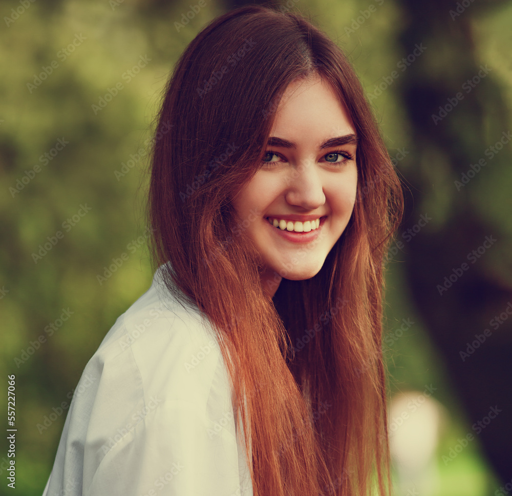Fototapeta premium Beautiful toothy smiling teen woman looking happy outdoors in white shirt on summer green trees background. Closeup portrait in bright sunny day. Toned