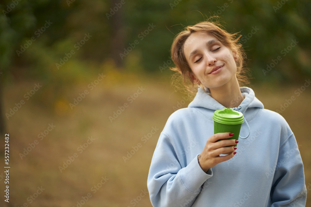 Satisfied cute caucasian girl in a blue hoodie keeps her eyes closed, smiles gently, holds takeaway coffee, likes to drink coffee from a takeaway cup. Smiling young girl holding a takeaway coffee. Satisfied cute caucasian girl in a blue hoodie keeps her eyes closed, smiles gently, holds takeaway coffee, likes to drink coffee from a takeaway cup. Smiling young girl holding a takeaway coffee.