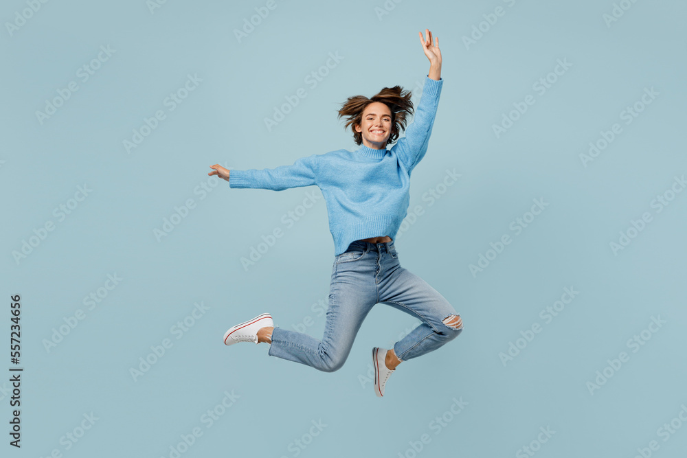 Full body overjoyed exultant young woman in knitted sweater look camera jump high with outstretched hands isolated on plain pastel light blue cyan background studio portrait People lifestyle concept
