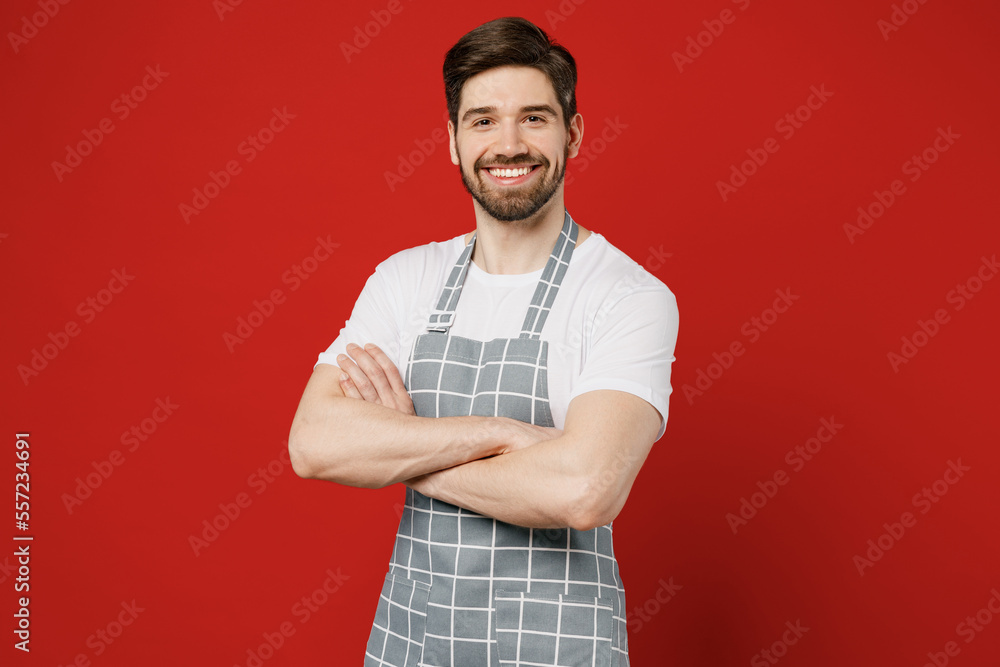 Young confident happy smiling fun male housewife housekeeper chef cook baker man in grey apron hold hands crossed folded look camera isolated on plain red color background studio Cooking food concept Young confident happy smiling fun male housewife housekeeper chef cook baker man in grey apron hold hands crossed folded look camera isolated on plain red color background studio Cooking food concept