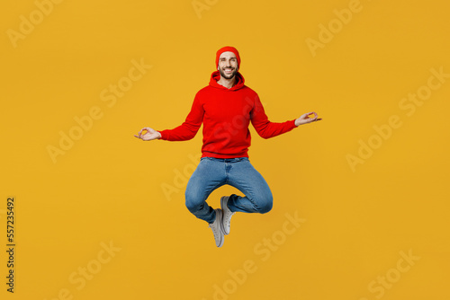 Full body young caucasian man wear red hoody hat look camera jump high hold spreading hands in yoga om aum gesture relax meditate try to calm down levitating isolated on plain yellow color background.