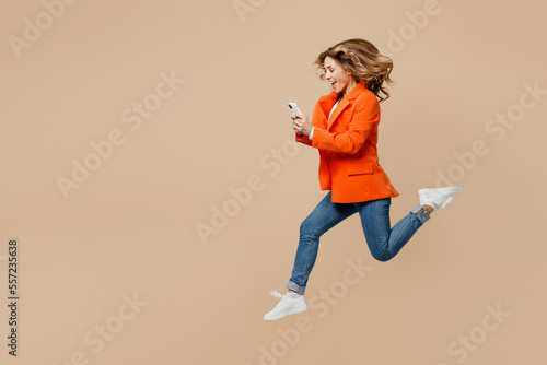 Full body side view young employee business woman corporate lawyer 30s wear classic formal orange suit glasses work in office jump high use mobile cell phone isolated on plain beige background studio.