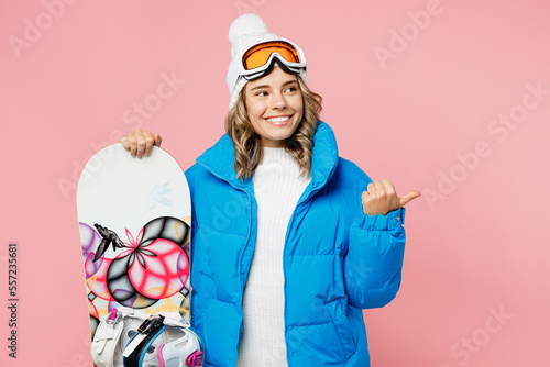 Snowboarder fun woman wear blue suit goggles mask hat ski padded jacket stand akimbo point aside on area isolated on plain pastel pink background Winter extreme sport hobby weekend trip relax concept