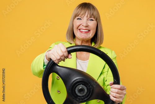 Elderly happy smiling woman 50s years old wearing green jacket white t-shirt hold steering wheel driving car look camera isolated on plain yellow background studio portrait. People lifestyle concept.