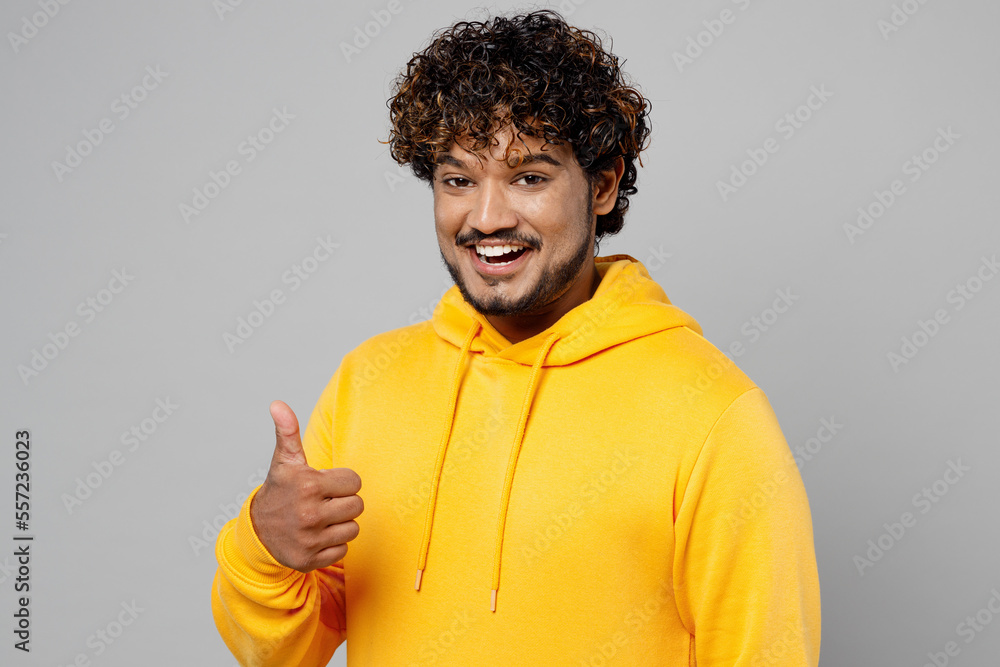 Young smiling happy fun cheerful satisfied cool Indian man 20s he wear casual yellow hoody showing thumb up like gesture isolated on plain grey background studio portrait. People lifestyle portrait. Young smiling happy fun cheerful satisfied cool Indian man 20s he wear casual yellow hoody showing thumb up like gesture isolated on plain grey background studio portrait. People lifestyle portrait.
