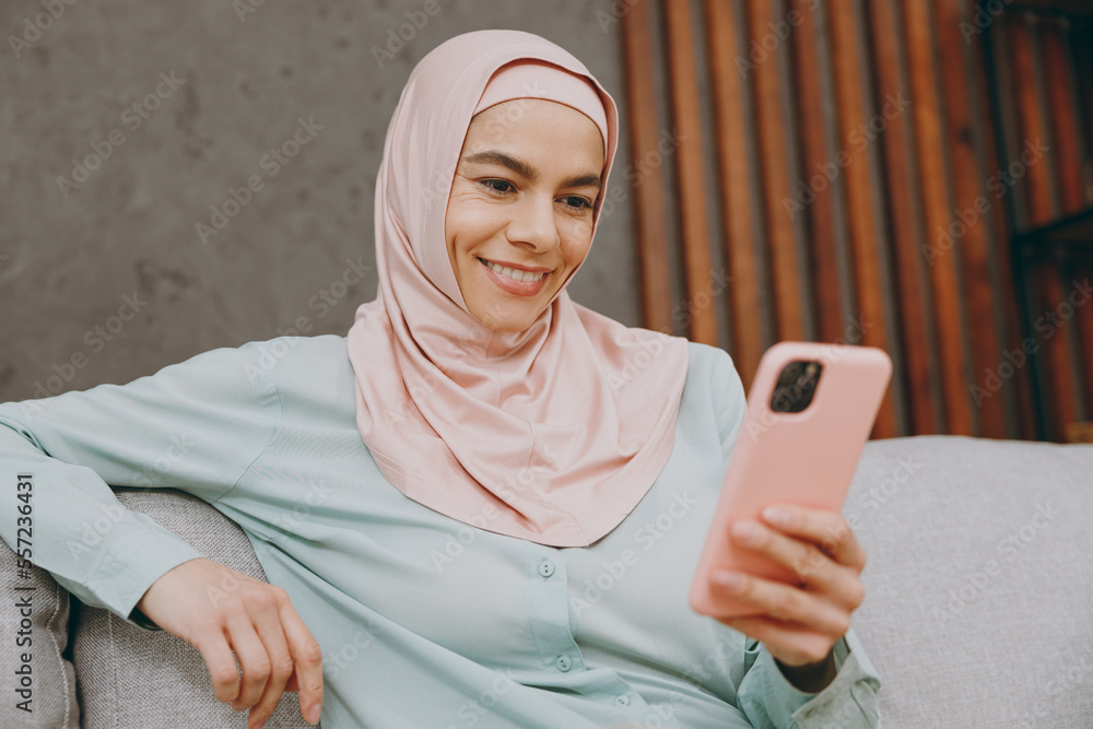 Young muslim woman wear hijab casual clothes use mobile cell phone sits on sofa couch stay at home flat rest relax spend free spare time in living room indoor. People uae middle eastern islam concept. Young muslim woman wear hijab casual clothes use mobile cell phone sits on sofa couch stay at home flat rest relax spend free spare time in living room indoor. People uae middle eastern islam concept.