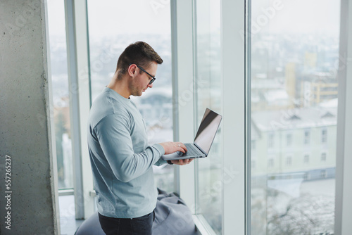Modern male manager in casual clothes stand by the window in the workspace with a laptop. Male freelancer works remotely.