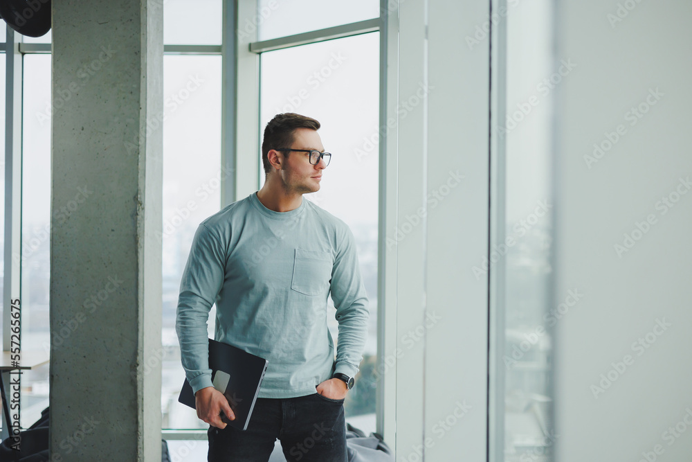 Modern male manager in casual clothes stand by the window in the workspace with a laptop. Male freelancer works remotely. Modern male manager in casual clothes stand by the window in the workspace with a laptop. Male freelancer works remotely.