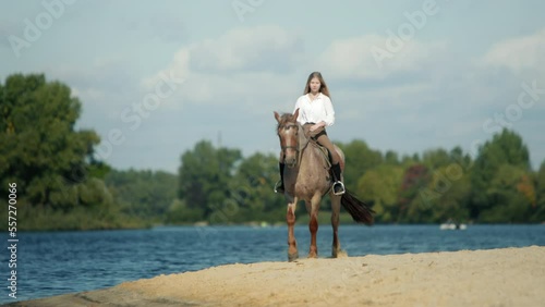 Equestrian rider walks her charming animal within sandy beach. Young, caucasian woman riding a horse along the shore with bluish lake water in the background. High quality 4k footage