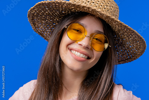 Charming woman in yellow eyewear and straw hat on blue studio backdrop. Smiling positive lady with asian appearance looking at camera. Summer, vacation, travel concept