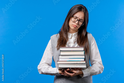 Lazy schoolgirl or student is dissatisfied with amount of books homework on blue background. Girl with long hair in displeasure, she is annoyed, discouraged frustrated by studies