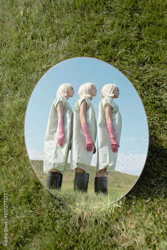 Conceptual photo of oval mirror lying on lawn reflecting alike young blond women in bizarre costumes standing in line against blue sky    photo