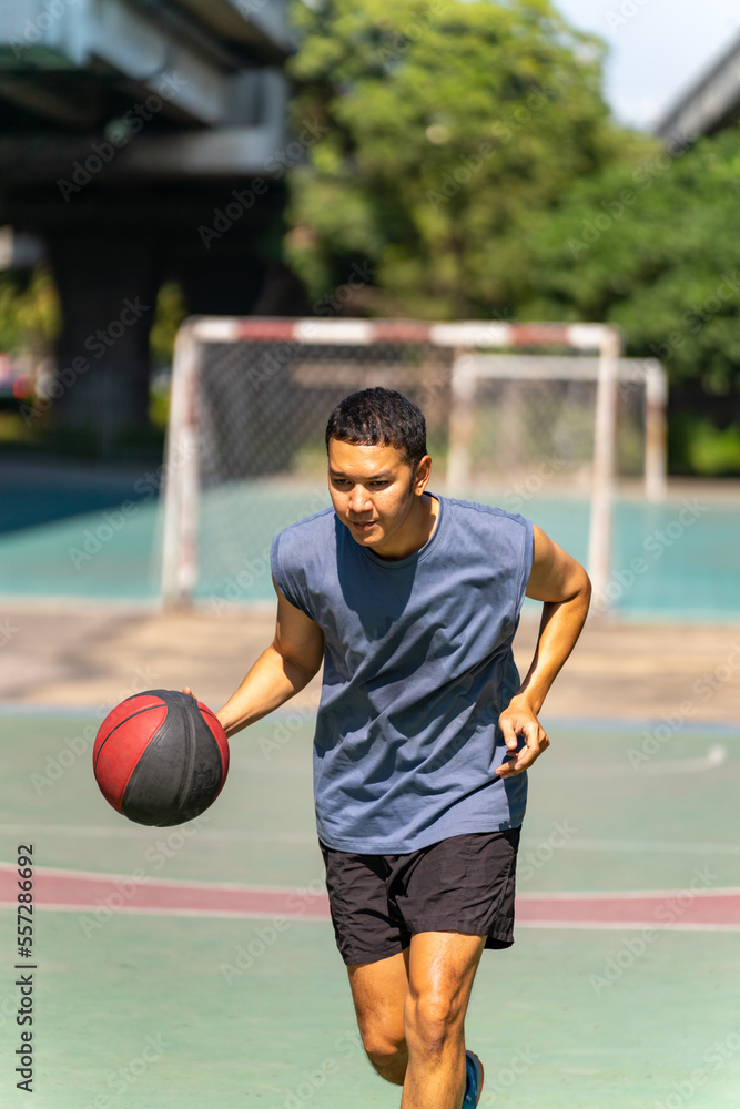 Naklejka premium Asian man basketball player practicing dribbling basketball on outdoors court under highway in the city. Sportsman do sport training workout exercise streetball at street court in summer sunny day.