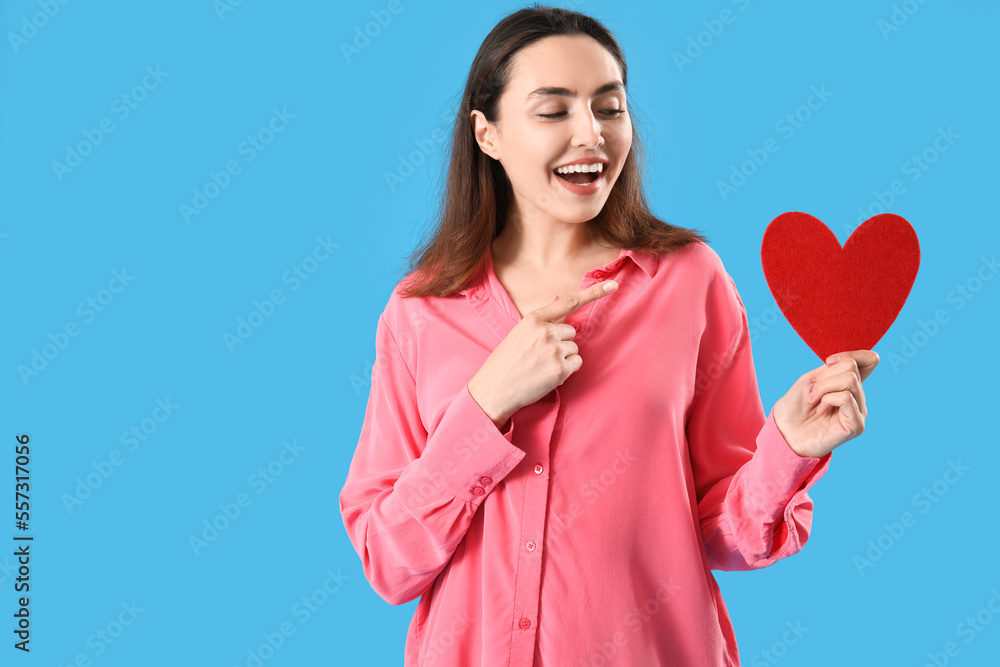 Young woman pointing at paper heart on blue background. Valentine's Day celebration Young woman pointing at paper heart on blue background. Valentine's Day celebration