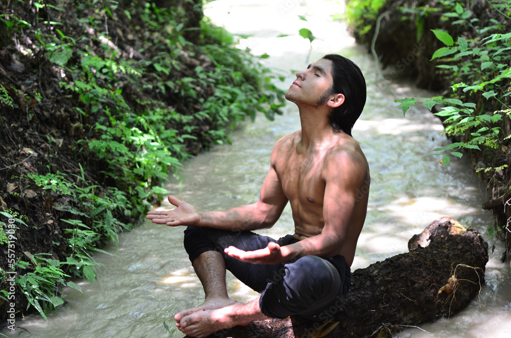 Joven hombre latino fornido meditando sobre un tronco en un arroyo o río en el bosque Joven hombre latino fornido meditando sobre un tronco en un arroyo o río en el bosque