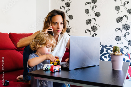 A single mother talks on the phone while working from home on her laptop and takes care of her young son, who plays with his toys. photo