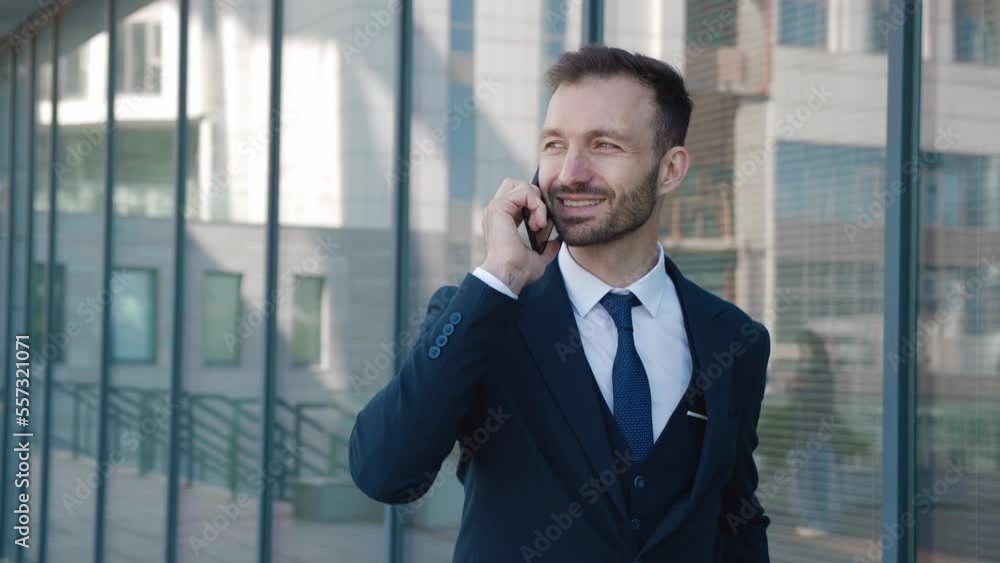 Smiling man entrepreneur talking mobile phone walking downtown. Cheerful entrepreneur in suit having phone call while walking outdoors in slow motion. Bearded male entrepreneur calling cell phone