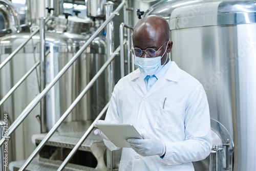 African American engineer in white coat working on digital tablet standing in workshop of factory