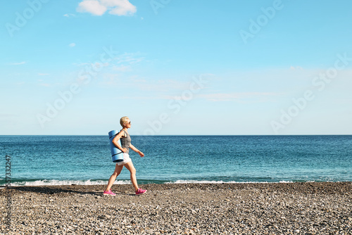Sporty blond woman in sport wear with yoga mat walking on the beach. Workout outdoors. Fitness, sport and healthy lifestyle concept.