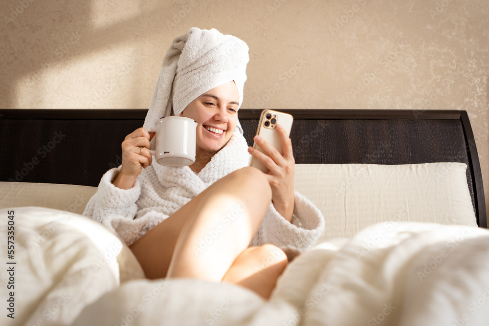 Sitting on a bed in a white dressing gown with a towel over her head is a woman with cup and mobile Sitting on a bed in a white dressing gown with a towel over her head is a woman with cup and mobile