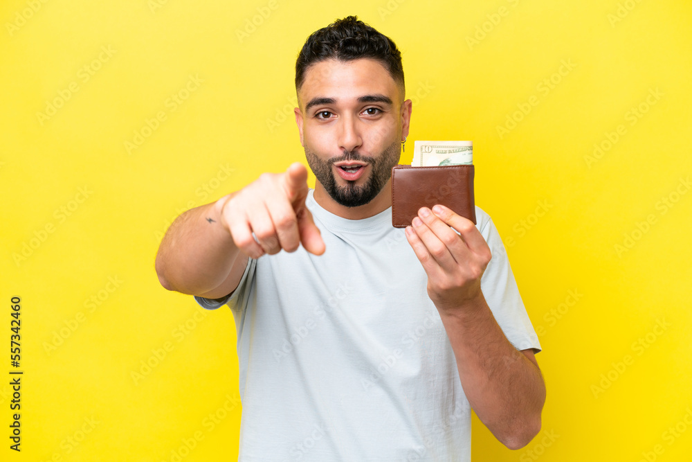 Young Arab man holding a wallet isolated on yellow background surprised and pointing front Young Arab man holding a wallet isolated on yellow background surprised and pointing front