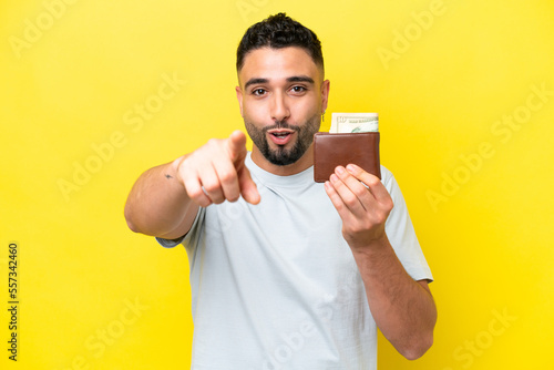 Young Arab man holding a wallet isolated on yellow background surprised and pointing front © luismolinero
