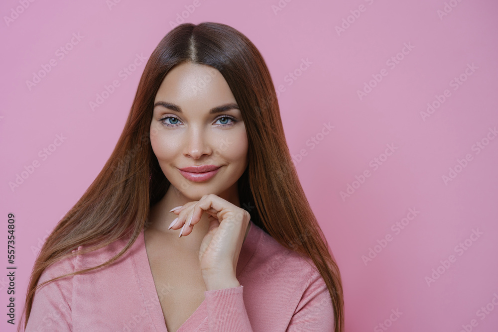 Pleasant looking brunette female fashion model keeps hand under chin, has full lips and applealing appearance, wears rosy jumper in one tone with background. Women, cosmetic and skin care concept Pleasant looking brunette female fashion model keeps hand under chin, has full lips and applealing appearance, wears rosy jumper in one tone with background. Women, cosmetic and skin care concept