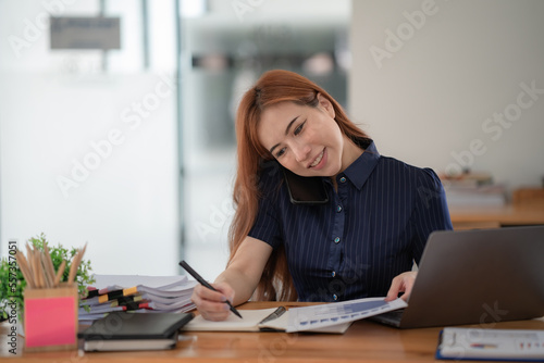 Young beautiful Asian woman using a smartphone and working with a laptop while sitting at an office desk, working from home concept.