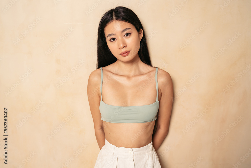 Fototapeta premium Beautiful asian girl in casual bra top posing on brown background with hands hidden behind her back