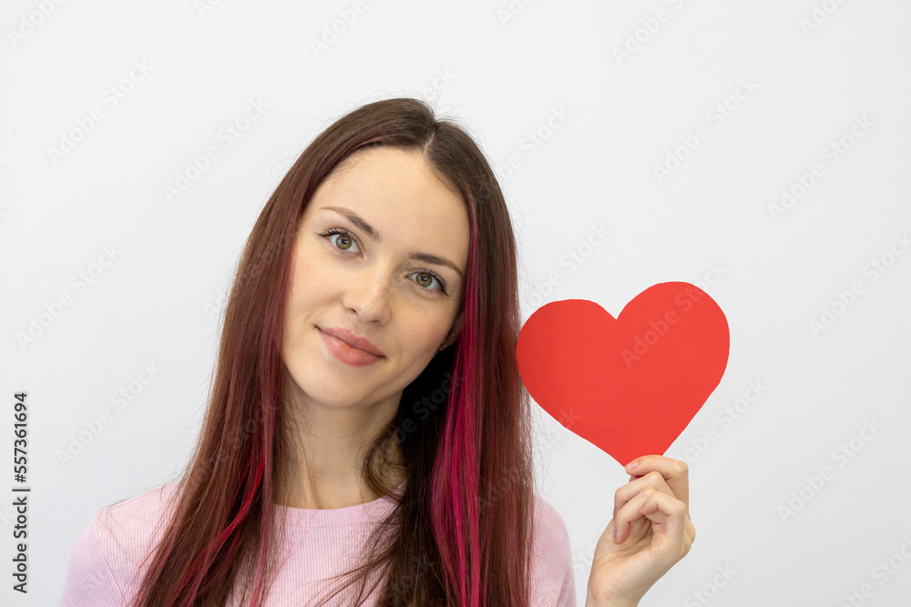 Beautiful young woman in love holding a red paper heart on a white background. The girl in pink looks straight at the camera and smiles Beautiful young woman in love holding a red paper heart on a white background. The girl in pink looks straight at the camera and smiles