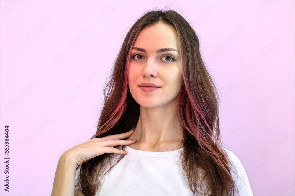 Young woman with a beautiful long neck wearing a white T-shirt against a glowing pink background. Curly girl with a fashionable hairstyle with pink strands of hair. Young woman with a beautiful long neck wearing a white T-shirt against a glowing pink background. Curly girl with a fashionable hairstyle with pink strands of hair.